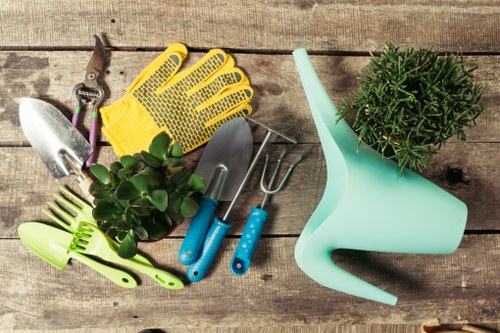 A collection of gardening tools and accessories arranged on a weathered wooden surface, including a pair of pruning shears, a yellow rubber glove, a small hand trowel, a hand rake, and a garden fork with blue handles, alongside a green watering can with a curved spout and a potted herb with dark green, needle-like foliage. The background reveals a natural outdoor setting, likely part of a landscaped garden near West Ham, featuring a mixture of textured wood and lush plant growth. The scene highlights essential gardening supplies used for planting, watering, and maintaining a garden space, suggesting preparation for outdoor garden care and landscaping tasks. The lighting appears natural, with soft shadows indicating a cloudy or partly sunny day, supporting the gardening theme associated with professional landscaping services provided by Landscapers West Ham.