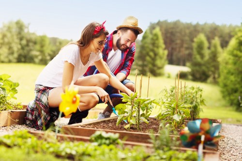 A couple is working together in a well-maintained garden located in West Ham, with a lush green background of trees and open fields. They are tending to raised garden beds filled with soil and young plants, possibly vegetable or flower seedlings, with the woman actively planting or watering and the man supervising or assisting. The garden features neatly edged borders, a small decorative bird figurine, and colorful garden ornaments. The outdoor space is illuminated by natural daylight, suggesting a clear, sunny day. Surrounding the garden are grassy areas and a spacious environment typical of suburban or semi-rural settings, reflecting professional landscaping and gardening efforts by Landscapers West Ham to enhance outdoor recreational or aesthetic spaces.