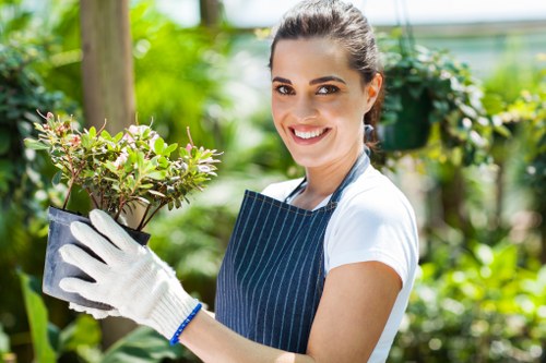 A young woman with dark hair tied back, wearing a white t-shirt, a blue striped apron, and gardening gloves, is standing in a lush green garden. She is smiling and holding a small potted shrub with pinkish-green leaves, clearly engaged in planting or caring for the plants. Behind her, there are various plants, small trees, and a wooden post, indicating a well-maintained outdoor space that could be part of a front or back garden in West Ham. The garden features a mix of densely packed foliage and open grassy areas, with natural sunlight illuminating the scene, suggesting a bright, pleasant day. The environment reflects a healthy, vibrant outdoor space, typical of residential gardening in East London, and aligns with professional landscaping and garden makeover services offered by companies such as Landscapers West Ham.