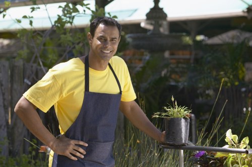 The image shows a smiling man in a yellow t-shirt and dark apron standing in a well-maintained garden in West Ham. He is holding a small black pot containing a young plant with thin green leaves, which appears to be in the process of planting or potting. The garden has a lush, green lawn with neatly trimmed grass, bordered by a variety of plants and shrubs. In the background, there is a wooden fence and a garden structure with a shaded area, possibly a pergola or a garden shed, partially obscured by foliage. The scene is lit by natural daylight, suggesting a clear, mild weather day. This outdoor space features a mix of paved and grassy surfaces, with some garden beds and planting areas visible. The presence of garden tools and the man's attire indicate active gardening work. The setting and activity reflect professional gardening and planting services commonly offered by Landscapers West Ham, supporting local outdoor maintenance and landscaping needs in the area.
