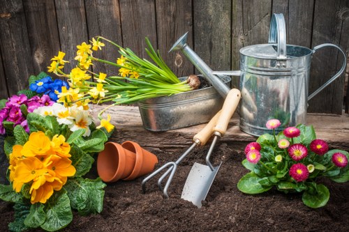 The image depicts a neatly prepared garden area with dark, well-tilled soil in the foreground, ready for planting or recent planting activity. On the left, there are colorful flowering plants with vibrant yellow, purple, and pink blossoms, supported by lush green leaves. In the middle, gardening tools including a hand rake and a small trowel are embedded in the soil, suggesting ongoing gardening work. Behind these tools, a row of empty terracotta plant pots is stacked, with some tipped over on the soil surface. To the right, a grey watering can and a metal container with a handle are positioned, indicating garden maintenance. The backdrop features a weathered wooden fence, providing a natural boundary for the garden space. The scene is illuminated by natural daylight, likely on a clear day, highlighting the rich earth tones and bright plant colours, evoking a professional gardening or landscaping setting typical of West Ham area gardens in London. Landscapers West Ham's services focus on outdoor maintenance and garden care, represented by the tools and plants in this landscaped outdoor space.
