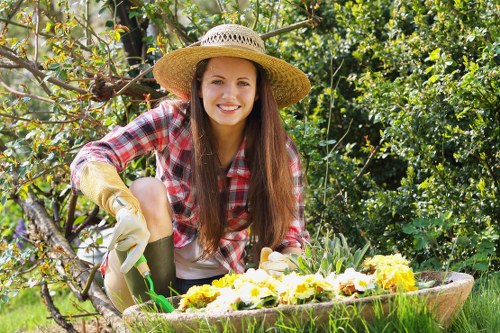 A young woman with long brown hair, wearing a wide-brimmed straw hat, a red and white checkered shirt, and gardening gloves, is kneeling in a lush garden. She is tending to a flower bed or preparing plants, with a smile on her face. The garden features a well-maintained lawn with dense green foliage and a tree branch nearby. Bright sunlight illuminates the scene, creating natural shadows and highlighting the vibrant greens of the plants and the yellow and white flowers in the flower bed. The environment suggests an outdoor space suitable for sustainable gardening practices, aligned with eco-friendly landscaping services offered by Landscapers West Ham in the local area. This scene reflects a peaceful, natural garden setting with a focus on plant care and outdoor maintenance in a suburban environment, possibly within the West Ham region or nearby postcode area.