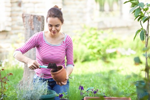 A young woman with dark hair tied back, wearing a long-sleeved pink and white striped top, is tending to her garden. She is holding a small terracotta pot in one hand and using a trowel with the other to plant or adjust soil inside the pot. The garden setting includes a lush, green lawn with various plants and flowers, some in pots and others growing directly in the soil. In the background, there are wooden elements, possibly a fence or garden furniture, and a tree trunk visible behind her. The environment appears to be well-maintained and vibrant, with natural daylight illuminating the scene, suggesting a pleasant, mild weather day. The overall space reflects a typical outdoor garden area suitable for gardening activities and outdoor leisure. The scene subtly supports gardening services related to planting and garden maintenance, fitting within the local West Ham area.