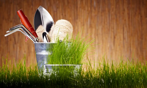 A close-up view of gardening tools and supplies placed on a grassy area of a garden, with a wooden fence in the background. The tools include a pair of hand gardening shears, a small garden fork, and a trowel, all with metal blades and red handles. There is also a ball of natural twine and a bunch of fresh green grass or young shoots in a metallic container. The scene highlights typical garden maintenance equipment set against a natural outdoor setting, suggesting preparation for planting or general garden care, aligned with landscaping services. The lighting indicates daytime with natural sunlight illuminating the scene, emphasizing the vibrant green of the grass and the metal surfaces of the tools. Such garden elements reflect the services offered by local West Ham landscapers specializing in gardening, lawn care, and outdoor maintenance within the area, contributing to lush and well-maintained outdoor spaces.
