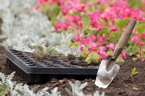 A gardening scene in a landscaped outdoor space, featuring a section of dark soil bordered by a flower bed with pink and white flowering plants. Nearby, a black seed tray sits on the soil with some seedlings or soil remnants visible inside. A small hand trowel with a wooden handle and metal blade is partially inserted into the soil, indicating active planting or soil preparation. The garden area appears well-maintained, with fresh soil and blossoming flowers, likely part of a garden makeover project undertaken by Landscapers West Ham, serving the West Ham area within London, focusing on gardening and outdoor improvements. The scene is captured under natural light, suggesting a clear day, with a tidy and inviting environment suitable for planting, lawn care, and landscape enhancement services.