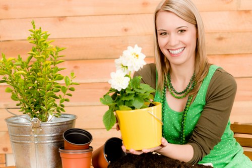 A woman with blonde hair, smiling, is seated outdoors in a garden, holding a yellow flower pot containing a plant with white flowers and green foliage. To her left, there is a metal bucket with a lush green shrub, and in front of her, stacked terracotta pots of various sizes are visible, suggesting gardening activity. The garden features a well-maintained lawn area with dense, lush grass, bordered by wooden decking or paving slabs, providing a neat outdoor setting. The background includes a wooden fence or wall, enhancing the garden's private and enclosed atmosphere. The scene appears to be lit with natural daylight, indicating a bright, possibly overcast day, suitable for outdoor gardening services offered by Landscapers West Ham in the local area. The overall environment showcases a tidy, organized, and inviting outdoor space focused on gardening and plant care, reflecting the company's expertise in landscaping and lawn maintenance in the West Ham region.
