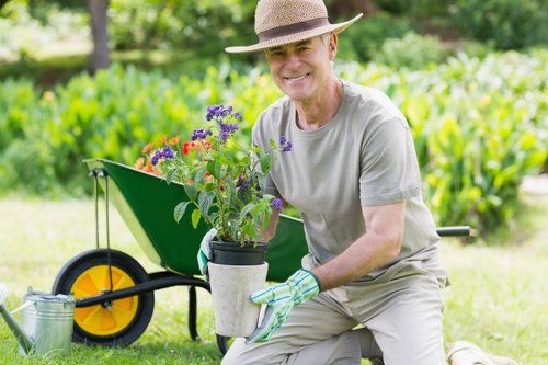 A gardener in a light grey t-shirt, wearing a straw hat and gardening gloves, is kneeling on a well-maintained lawn in a back garden in West Ham, holding a potted flowering plant with purple, red, and pink blooms. Behind him, a green wheelbarrow with a yellow wheel contains additional gardening tools and plants. The garden features a neatly clipped hedge line, a flower bed with a curved border, and a variety of lush green shrubs and trees in the background, suggesting a healthy, natural outdoor environment. The overall scene is set on a bright, sunny day with natural light illuminating the vibrant greens and colourful flowers, demonstrating typical garden maintenance activities supported by professional landscaping and gardening services such as those provided by Landscapers West Ham.
