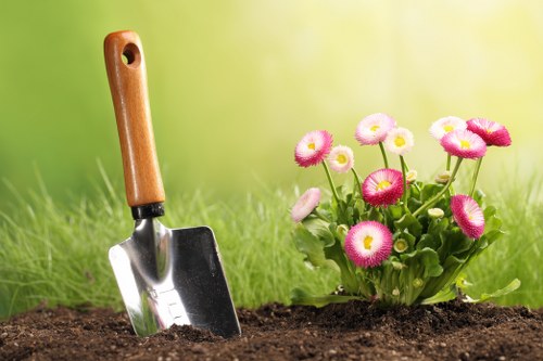 A close-up image of a garden bed showing dark, moist soil prepared for planting. In the foreground, a small trowel with a wooden handle and metal blade is partially inserted into the soil, suggesting recent gardening activity. To the right, there is a vibrant cluster of pink and white primrose flowers with rounded petals and bright green leaves, adding colour to the landscaped outdoor space. The background features a blurred green lawn with freshly mown grass, indicating well-maintained grounds typical of a residential garden in West Ham. The scene is illuminated by natural daylight, possibly during a mild, cloudy morning or afternoon, creating a soft, inviting atmosphere. This setting reflects professional gardening and landscaping services that include planting, soil preparation, and maintenance of flower beds within a suburban garden environment in East London, near postcode regions such as E13 or E6, consistent with services offered by Landscapers West Ham.