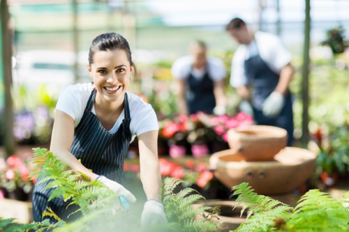 A young woman with dark hair tied back, wearing a white t-shirt and a blue apron, is smiling at the camera while working in a greenhouse or garden centre, surrounded by various potted plants and flowers. She appears to be engaged in gardening or plant care activities, with her hands possibly resting on or near the plants in front of her. The background shows blurred individuals, likely staff or customers, wearing similar aprons and handling plants, indicating an outdoor or greenhouse setting focused on gardening services in West Ham or nearby areas. Natural daylight filters through the greenhouse structure, illuminating the lush greenery and vibrant pink and red flowers, as well as the textured soil and plant pots. The scene highlights a professional gardening environment characteristic of Landscapers West Ham, emphasizing plant care, gardening work, and outdoor landscaping maintenance in a well-kept, green outdoor space or garden centre.